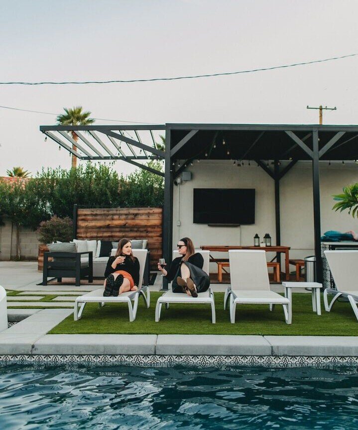 Two women lounge on poolside chairs, chatting and holding drinks in a backyard with a covered patio and outdoor seating behind them.