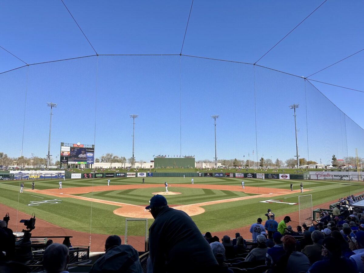 Baseball stadium during a game, players on the diamond with protective netting in the foreground and a clear blue sky above.